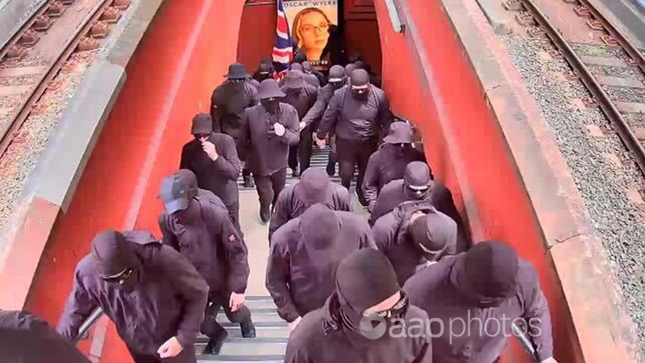 About 20 men all dressed in black walk up stairs to a train platform  